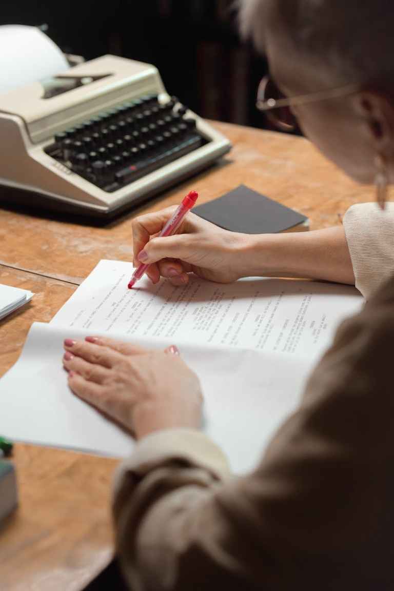 A woman reviews and edits papers using a red pen near a vintage typewriter..jpg